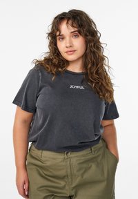 Sieviete ar cirtainiem brūniem matiem, valkā izbalējušu melnu T-kreklu ar uzrakstu "JOYFUL" un olīvu zaļas krāsas bikses, stāvot ar vienu roku kabatā.