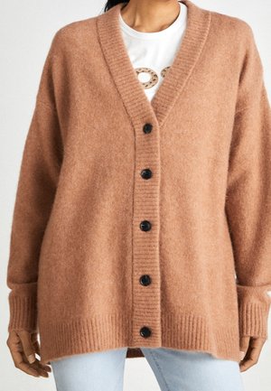 Vrouw draagt een loszittend, zacht bruin vest met knopen over een wit shirt met een afbeelding, gecombineerd met lichtblauwe spijkerbroek.