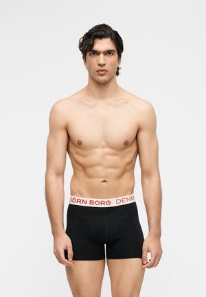 Muž bez trička s vyrysovanou muskulatúrou, oblečený v čierne boxerky s bielym pásom označeným "BJÖRN BORG DENMARK", stojaci pred jednoduchým pozadím.