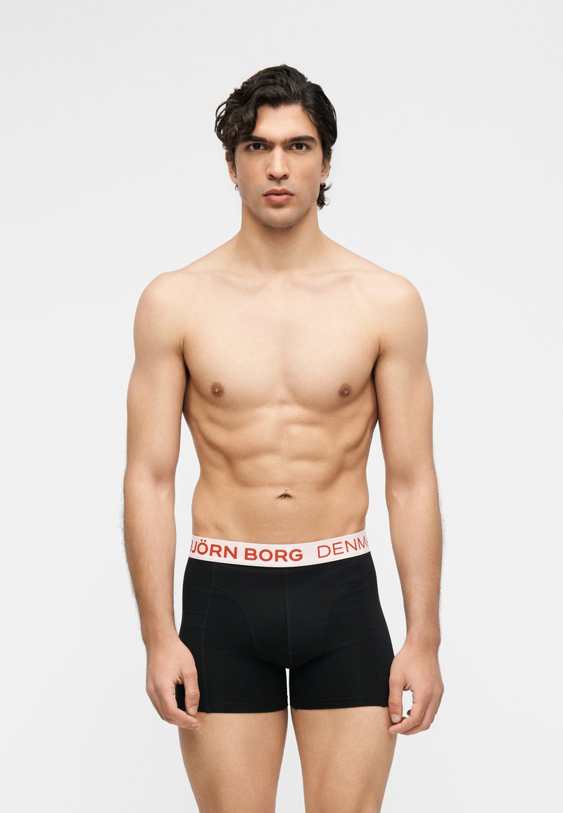 Άνδρας χωρίς πουκάμισο με μυώδη σωματότυπο, φοράει μαύρα μποξεράκια με λευκή ζώνη που γράφει "BJÖRN BORG DENMARK" και στέκεται μπροστά από μια απλή φόντο.