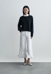 Camisola preta de malha com bainha canelada, combinada com jeans brancos de perna larga e barra dobrada. Saltos em tom de branco sujo com bico pontiagudo completam o conjunto.