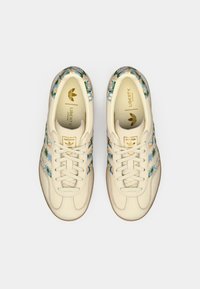 Sapatilhas de couro brancas com detalhes florais azuis, três riscas distintivas e uma sola de borracha texturizada. Detalhes do logo em dourado na língua.