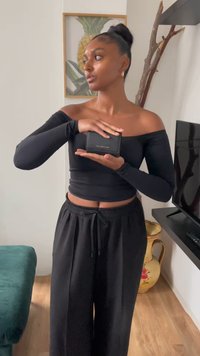 Portafoglio in pelle nera con lettering dorato, tenuto di fronte a una figura vestita con un top nero scollato e pantaloni neri, ambientato all'interno.