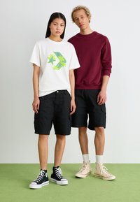 To personer står på grønne gulve. Den ene har en hvid t-shirt med et neongrønt grafik og sorte cargo shorts. Den anden er iført en bordeaux sweatshirt med sorte shorts, kombineret med beige og sorte sneakers.