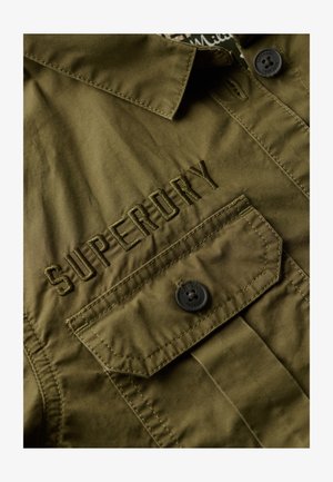 Camisa verde oliva con cuello abotonado, que presenta el logotipo bordado "SUPERDRY" en el pecho y un bolsillo con un botón negro. Textura suave.