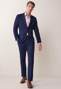 Traje azul marino con un corte ajustado, que incluye un blazer de un botón, combinado con una camisa rosa claro y zapatos brogue de cuero marrón.