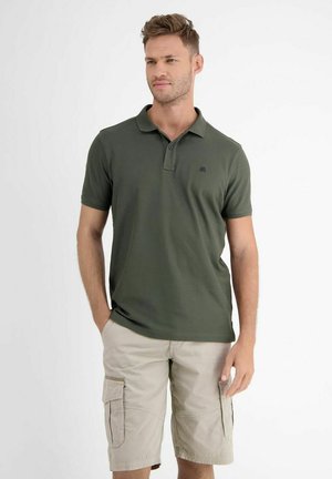 Mann trägt dunkelgrünes Polohemd und beige Cargoshorts, steht mit einer Hand in der Tasche, schaut leicht nach rechts, einfarbiger weißer Hintergrund.