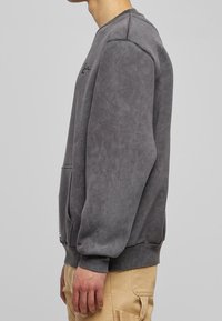 Šedá mikina s kulatým výstřihem vyrobená z měkké, texturované tkaniny. Obsahuje přední kapsu a decentní výšivku loga. Volný střih s žebrovanými manžetami.