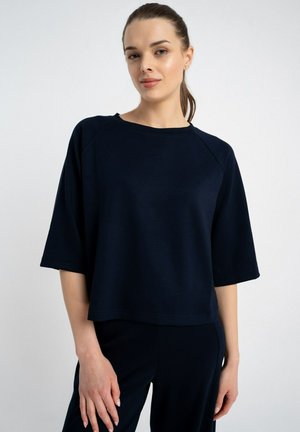 Kobieta z upiętymi włosami, ubrana w luźną granatową bluzkę z krótkim rękawem i pasujące spodnie, stojąca na jednolitym tle.