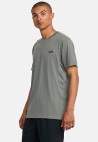 Camiseta atlética gris de manga corta, con cuello redondo y un pequeño logo negro en el pecho. Presenta un panel lateral texturizado para mejorar la transpirabilidad.