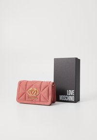 Sarkana zamšas maks ar quilted rakstu, zelta aksesuāriem un logotipu. Iesprausts melnā kastē ar uzrakstu "LOVE MOSCHINO" baltā krāsā.