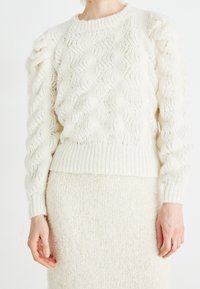 Pull blanc en maille torsadée avec motif zigzag texturé et ourlet côtelé. Manches bouffantes. Porté avec une jupe assortie de couleur crème.