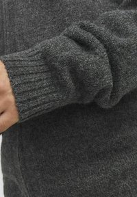 Grauer Strickpullover mit geripptem Saum, weicher Textur und lockerer Passform. Eine Nahaufnahme zeigt die Details des Stoffes und die Farbvariationen.