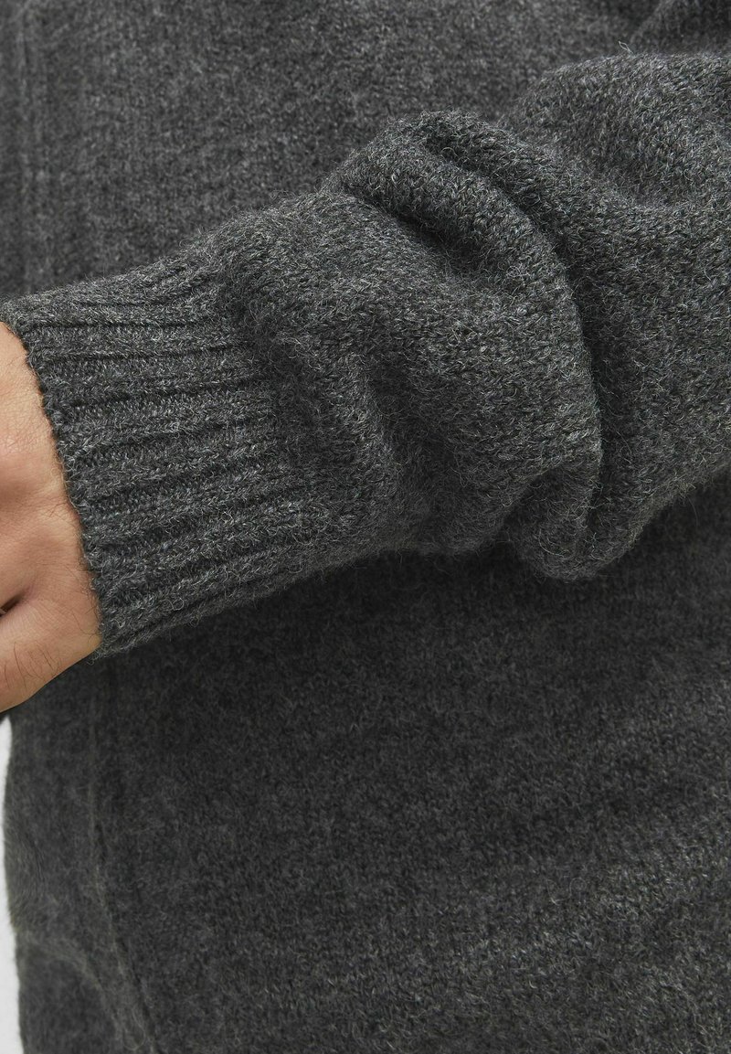 Grauer Strickpullover mit geripptem Saum, weicher Textur und lockerer Passform. Eine Nahaufnahme zeigt die Details des Stoffes und die Farbvariationen.