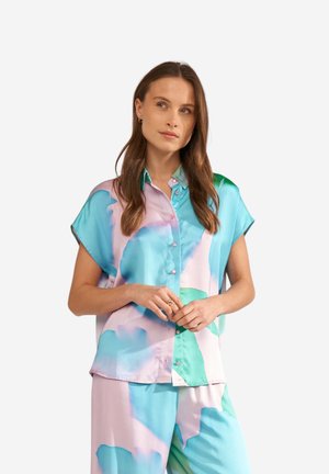 Žena oblečená v voľnej pastelovej košeli s krátkym rukávom na gombíky a v zodpovedajúcich nohaviciach s abstraktnými modrými, zelenými a ružovými vzormi, stojaca so založenými rukami.
