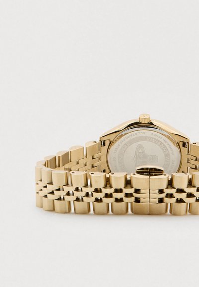 Zelta metāla saite pulksteņa siksniņai ar atvērtu aizdari, redzama pulksteņa korpusa aizmugure ar iegravētiem zīmola un tehniskajiem datiem.