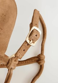 Hnedý semišový remienok sandálov so zlatým kovovým prackou a nastaviteľnými dierkami, upevnený uzlami na neutrálom pozadí.