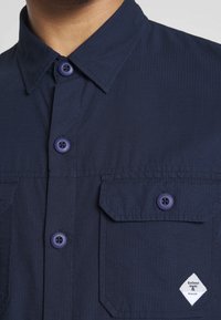 Camicia a maniche corte blu navy in tessuto strutturato, con due tasche sul petto, bottoni viola e una toppa con logo a forma di diamante sul basso a destra.