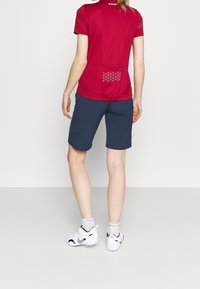 Červená krátká tričko s krátkým rukávem a zadní kapsou se geometrickým vzorem, v kombinaci s námořnickými modrými šortkami. Oblečeno s bílými sportovními botami a ponožkami.