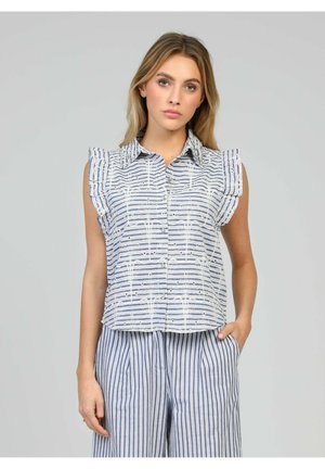 Femme portant un haut sans manches boutonné avec des épaules à volants et un pantalon à jambes larges rayé bleu et blanc, debout devant un fond uni.