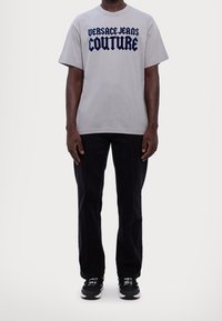 Uomo che indossa una maglietta grigia "Versace Jeans Couture", pantaloni neri con cuciture anteriori e scarpe da ginnastica nere con suole bianche e lacci con logo.