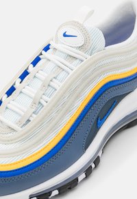 Nike Air Max 97 tenisky s vrstveným designem, kombinující síťovinu a kůži v modré, šedé, bílé a žluté. Silná černá podrážka s viditelnou vzduchovou jednotkou.
