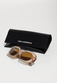 Karl Lagerfeld solbriller med en lys rosa gjennomsiktig ramme og brune tonede glass, akkompagnert av et svart etui med hvitt logo.