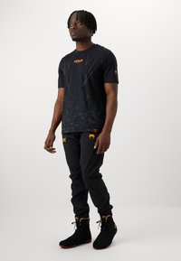 Camiseta deportiva negra con patrón de camuflaje y logo "Venum" en naranja, combinada con pantalones negros que presentan el logo "UFC" en amarillo, y zapatillas deportivas negras.