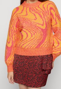 Kvinna klädd i en ljus orange och rosa virveltröja som matchar med en svart minikjol med ett rött hjärt-mönster och sidofrill. 