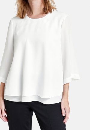 Blouse blanche à couches avec un col rond et des manches trois-quarts. Fabriquée en tissu léger, elle présente une texture lisse et un éclat subtil.