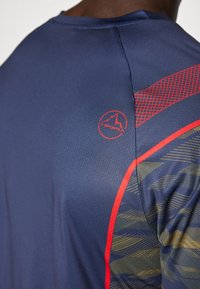 Granatowa koszulka sportowa wykonana z teksturowanej tkaniny, z czerwono-zielonymi ukośnymi paskami i okrągłym dekoltem z logo na ramieniu.