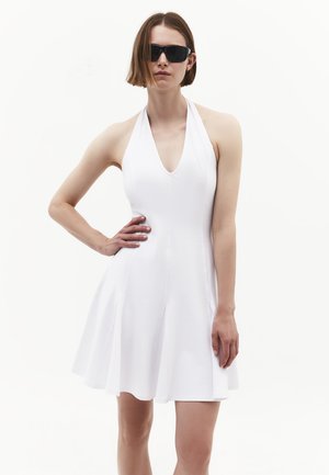 Donna con occhiali da sole neri e abito bianco senza maniche con collo all'americana, in piedi con una mano sul fianco su sfondo bianco.