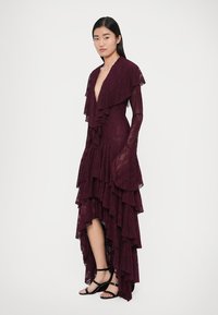 Vestido de renda cor vinho com folhos em camadas, mangas compridas, decote em V profundo e bainha assimétrica. Apresenta textura transparente e design fluido.