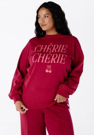 Vrouw met lang, donker haar die een bordeauxrode sweatshirt en broek draagt, de sweatshirt heeft de tekst "CHÉRIE CHÉRIE" met daaronder een kersgrafiek.