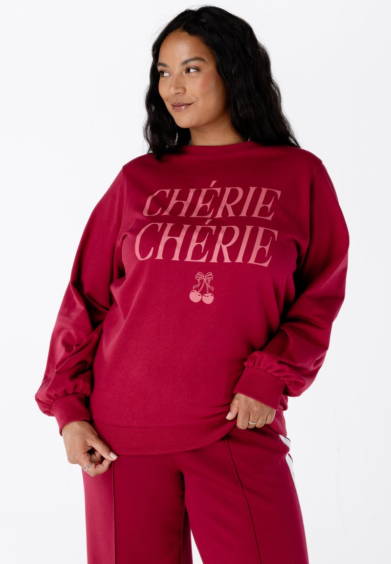 Femme avec de longs cheveux sombres portant un sweat-shirt bordeaux et un pantalon, le sweat-shirt arbore l'inscription "CHÉRIE CHÉRIE" avec un graphique de cerise en dessous du texte.