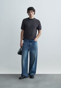 Black patterned t-shirt with short sleeves, paired with wide-leg blue denim jeans. Model holds a black bag and wears black shoes.