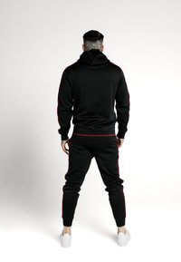 Tuta nera con strisce laterali rosse. Presenta un cappuccio e gambe affusolate. Realizzata in tessuto liscio, completata con scarpe da ginnastica bianche.