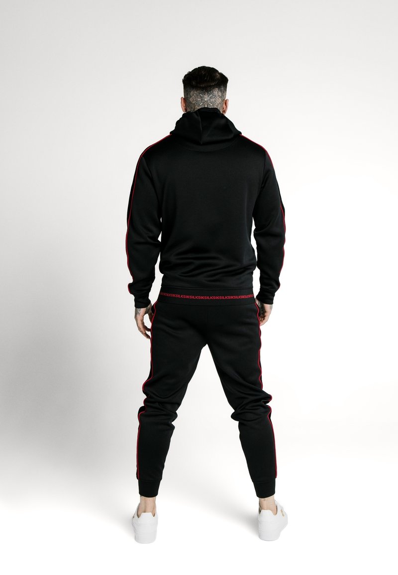 Tuta nera con strisce laterali rosse. Presenta un cappuccio e gambe affusolate. Realizzata in tessuto liscio, completata con scarpe da ginnastica bianche.