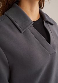 Primer plano de una persona que lleva una chaqueta de cuello oscura sobre una camisa negra, mostrando el cuello y la parte superior del pecho.