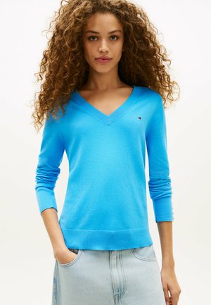 Vrouw met krullend haar die een felblauwe V-hals trui en een lichte denim rok draagt, staand met één hand in haar zak.