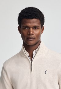 Maglione zip-up color beige chiaro con colletto a coste e maniche lunghe, con un piccolo logo blu navale sul lato sinistro del petto. Tessuto morbido e texturizzato.