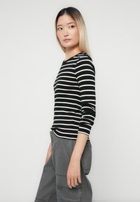 Woman with straight ombre hair wearing a black and white striped long-sleeve shirt and gray cargo pants, standing sideways against a white background.