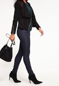 Schwarze Wildlederjacke mit asymmetrischem Design, grünes Oberteil darunter, blaue Skinny-Jeans und schwarze Ankle-Boots mit spitzem Zehenbereich. Schwarze Handtasche in der Hand gehalten.