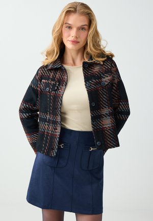 Femme aux cheveux blonds ondulés portant une veste à carreaux noir et rouge, un top beige et une jupe bleu marine avec poches avant, debout devant un fond uni.