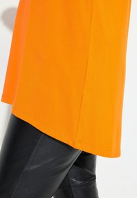 Oranges Baumwoll-T-Shirt mit geradem Saum, kombiniert mit schwarzen Leggings aus Kunstleder mit glänzender Textur und figurbetontem Design.