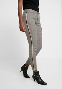 Pantalons à carreaux beiges et noirs avec une coupe slim, dotés de bandes latérales rouges et blanches. Portés avec des bottines noires.