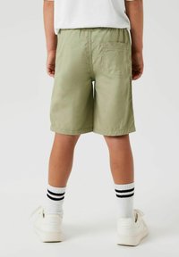 Shorts en coton vert clair avec une taille élastique et une poche arrière, portés avec des baskets blanches et des chaussettes à rayures noires, vus de derrière.