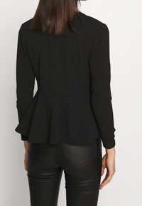 Schwarze Peplum-Jacke mit langen Ärmeln, tailliertem Oberteil und strukturierter Stoff, kombiniert mit schwarzen, enganliegenden Hosen. Maßgeschneiderte Details am Rücken.