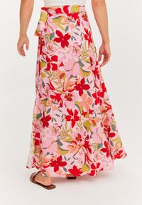 Žena v dlhých ružových sukňach s červeným, oranžovým a zeleným kvetinovým vzorom a hnedými plochými sandálmi, z pohľadu zozadu.