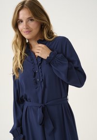 Robe bleu marine en tissu doux, dotée d'un col volanté, d'une patte de boutonnage et de manches longues avec des poignets ajustés. Comprend une ceinture à nouer à la taille.